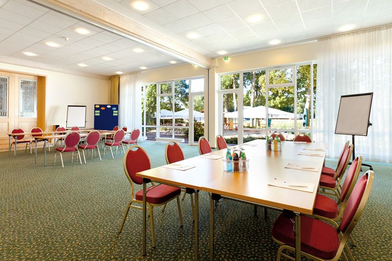 Lichtdurchfluteter Konferenzraum mit Terrasse Konferenzraum mit Tischen und roten Stühlen, bereit für ein Meeting, mit Blick auf eine Terrasse durch große Fenster.